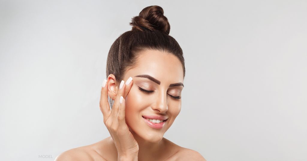 Woman with brown hair up in a bun is smiling and touching her face with her hand. (model)