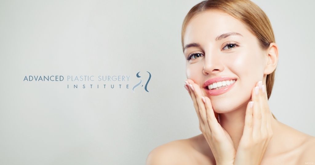 A blond woman smiling and gently touching her face.  (MODEL) The Advanced Plastic Surgery Institute logo.