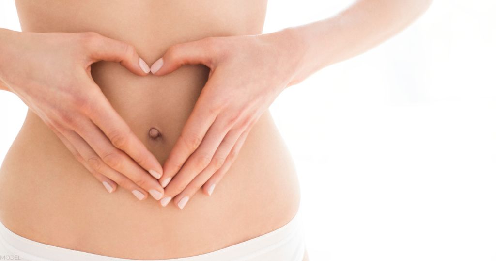 Woman with hands in heart shape around navel (MODEL).