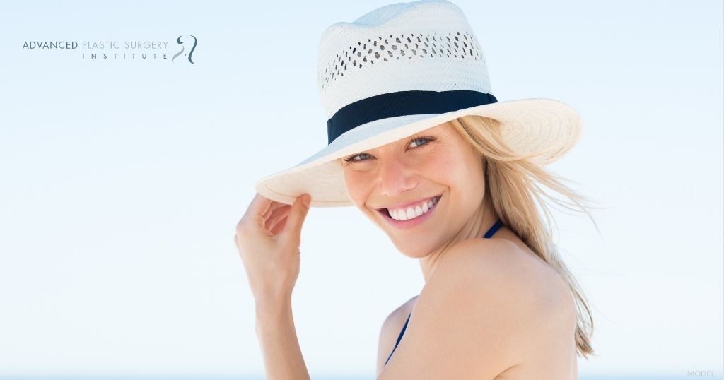 Young woman wearing a hat and smiling at the beach (MODEL) Advanced Plastic Surgery Institute Logo