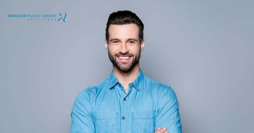 A man in a blue shirt standing in front of a gray background (MODEL) Advanced Plastic Surgery Institute Logo